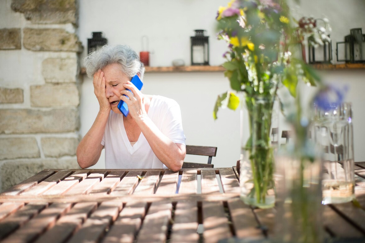 Distressed woman in summer kitchen talking on phone, sunlight and flowers in background — showing hidden signs of summer anxiety