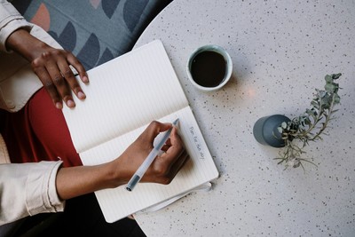 Person sitting at a table writing in a journal with a cup of black coffee nearby, focusing on hands only, representing cognitive behavioral therapy and self-reflection.