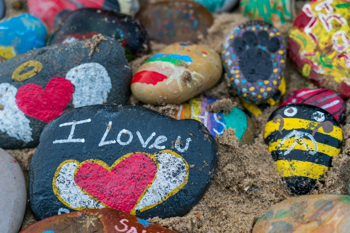 Painted rocks on sand, including designs like a bee, a heart with wings, a footprint, and the message “I love you”—part of a calming summer art activity for stress relief and creative expression.