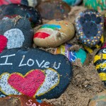Painted rocks on sand, including designs like a bee, a heart with wings, a footprint, and the message “I love you”—part of a calming summer art activity for stress relief and creative expression.