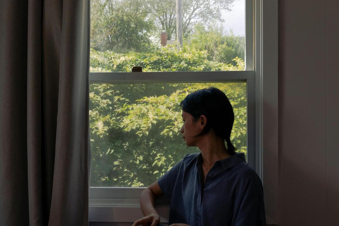 Woman standing indoors in a dimly lit room, looking out at a bright summer day, experiencing mid-summer sadness as she reflects on the feeling that summer is slipping away.