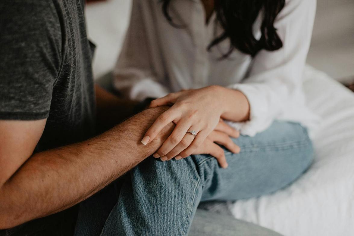 A man sits beside a woman on a bed, resting his hand on her leg as she places her hand on his, showing care and support during a difficult moment.