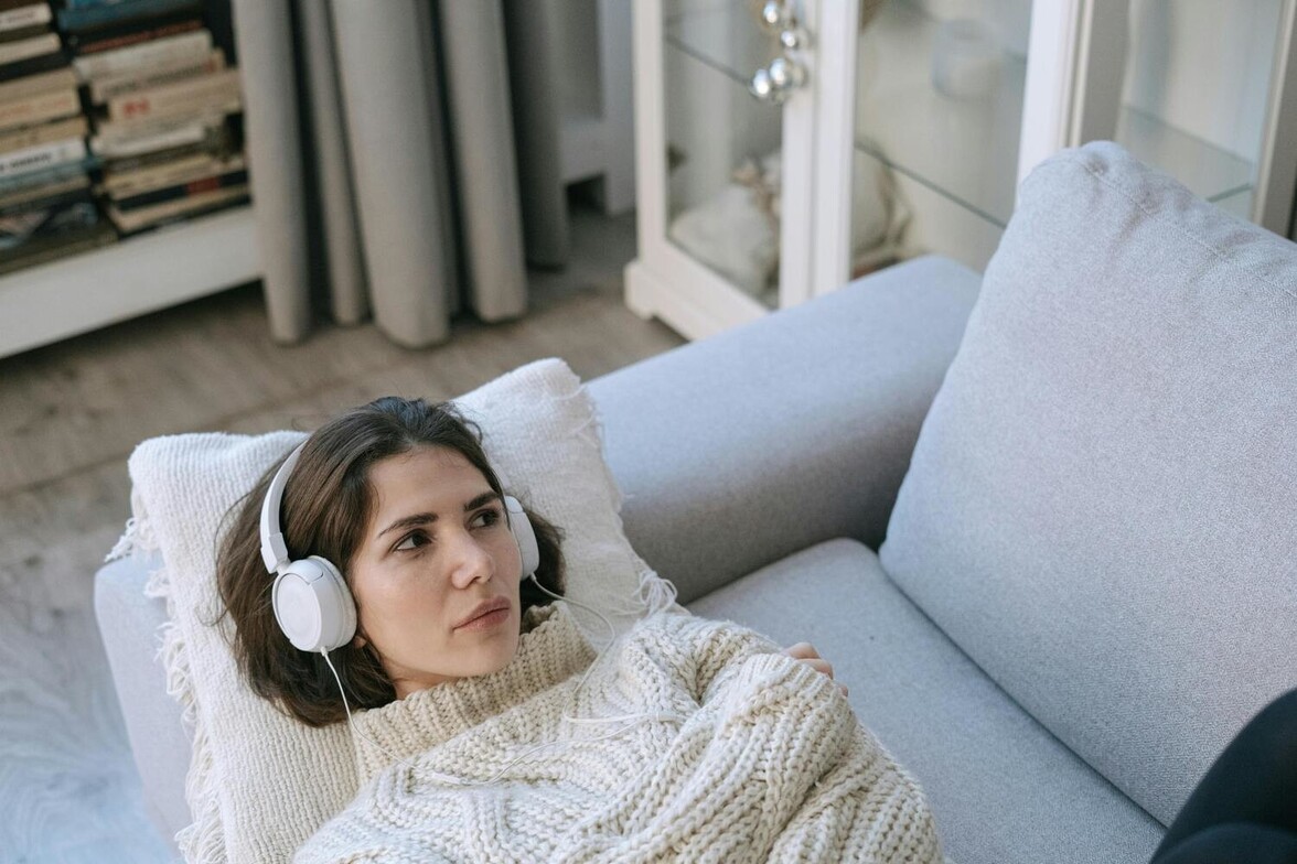 Woman lying on a couch in a bright room, wearing headphones and a heavy sweater, with a sad and distant expression.