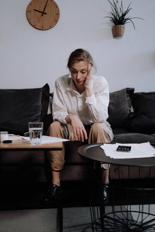 Woman sitting on a couch with her hand on her face, looking down at piles of papers — representing overwhelm, stress, or symptoms of depression.