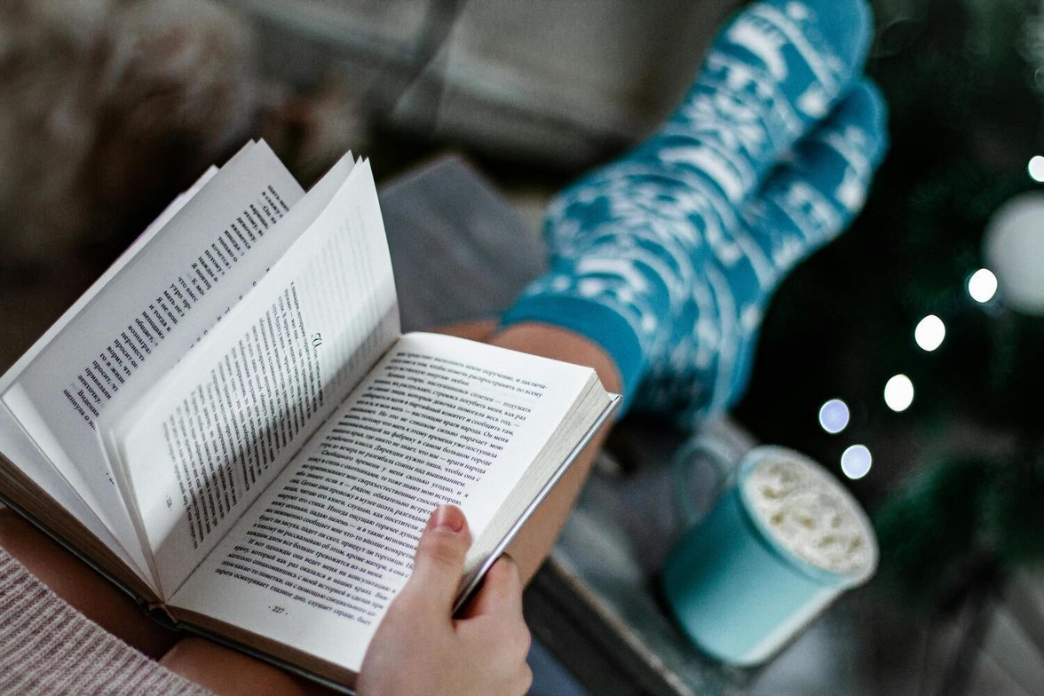 Woman sitting in a cozy chair wearing patterned winter socks, reading a book and enjoying a quiet moment to rest during the winter break.