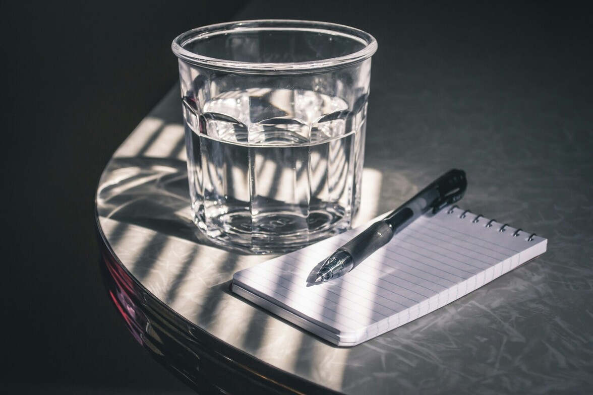 A glass of water beside an open mini memo pad and pen, representing reflection, small daily actions, and mindful planning.